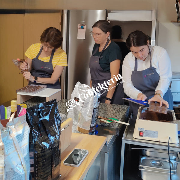 Drei Frauen stehen an der Pralinenstraße, eine füllt die Pralinenformen mit Kuvertüre, die zweite hält eine Form über das Warmhaltebecken und lässt die überflüssige Kuvertüre aus der Form fließen.