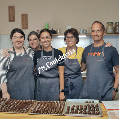 fünf glückliche Menschen stehen in einer Backstube, auf dem Tisch vor ihnen liegen hunderte glänzende Pralinen. Familie Bauer hat diese Pralinen während eines Pralinenkurses selbst hergestellt